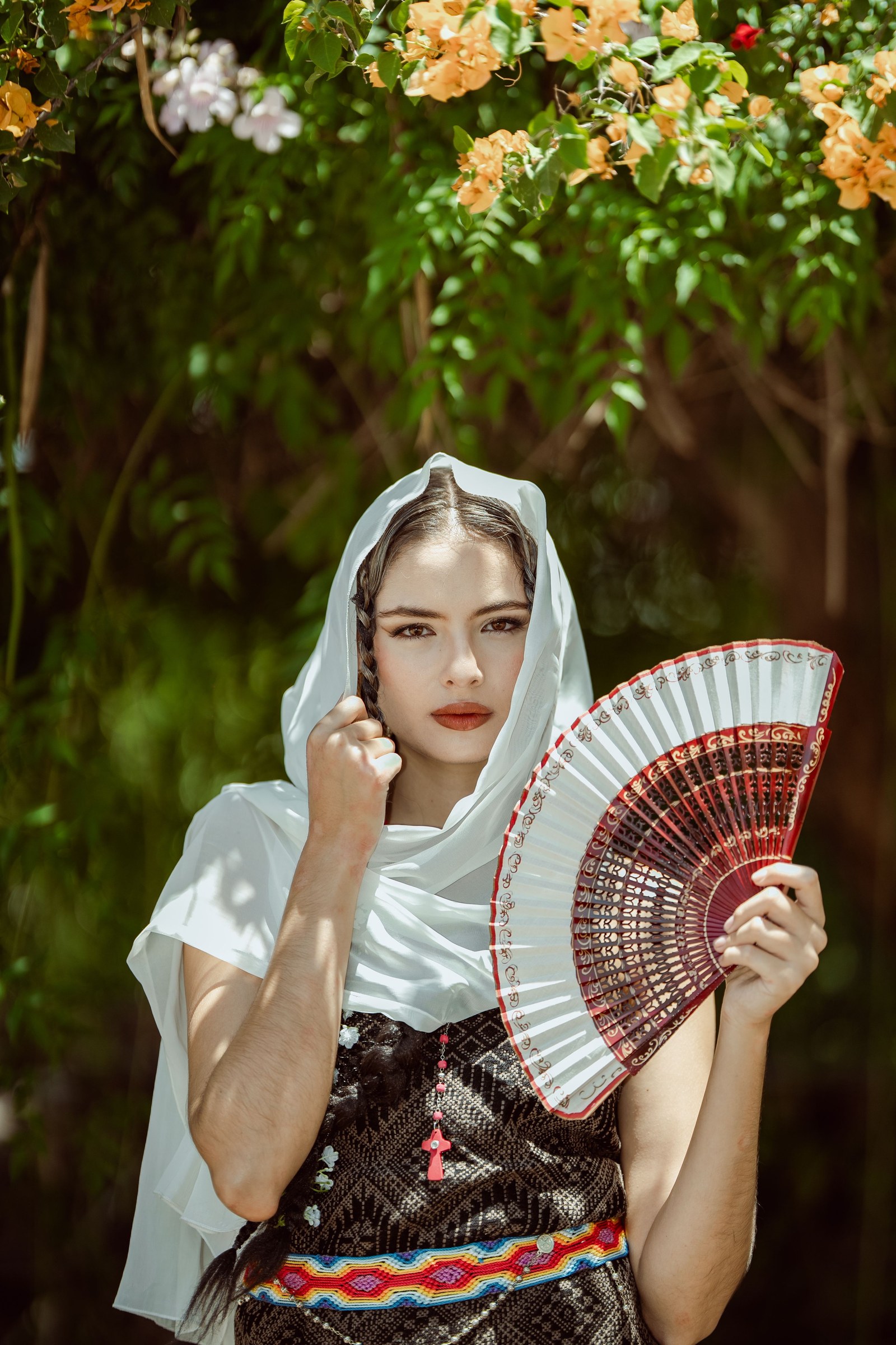 A woman in traditional Mexican dress holding a decorative fan beneath colorful flowers.