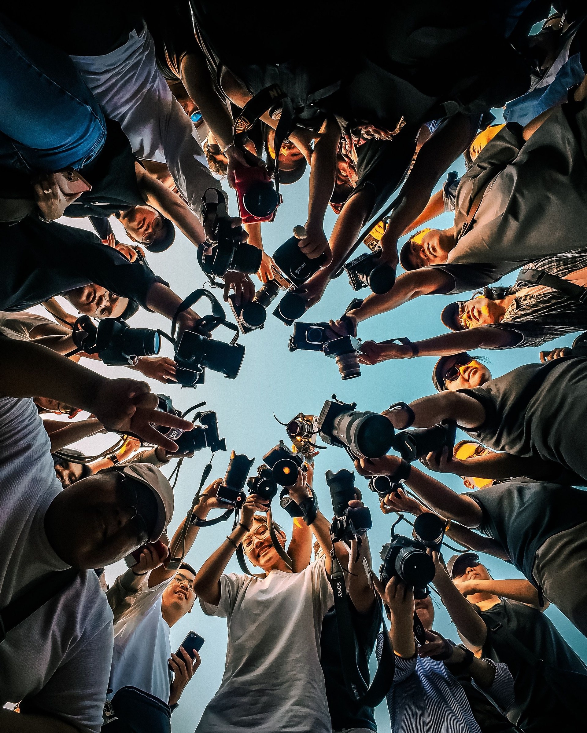 A circle of photographers pointing their cameras toward the center, viewed from below against a blue sky.
